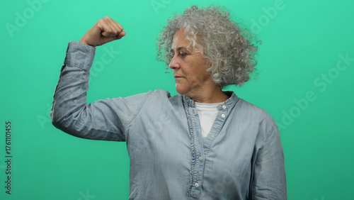 Canvas Print Elderly woman with grey hair does a strength gesture against an isolated green background wall, embodying empowerment and resilience