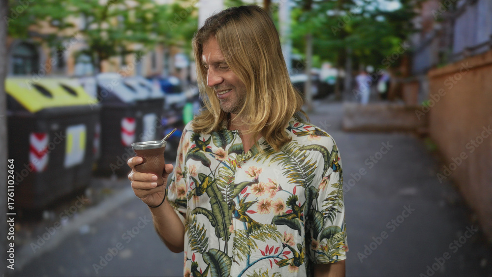 Fototapeta premium Man with long blond hair smiles while holding a traditional mate cup on a cobblestone street in sunlight; serenity.