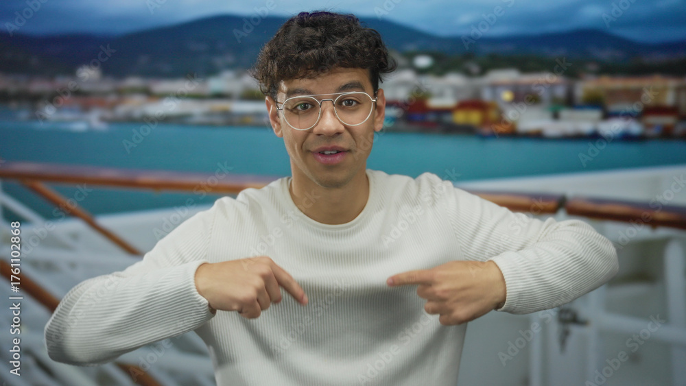 Fototapeta premium Young hispanic man wearing glasses and sweater waving hand on cruise deck with railing and sea in view; adventure greeting travel excitement.