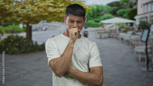 Man with hand to mouth coughing at outdoor restaurant terrace street scene; health concern and vulnerability.