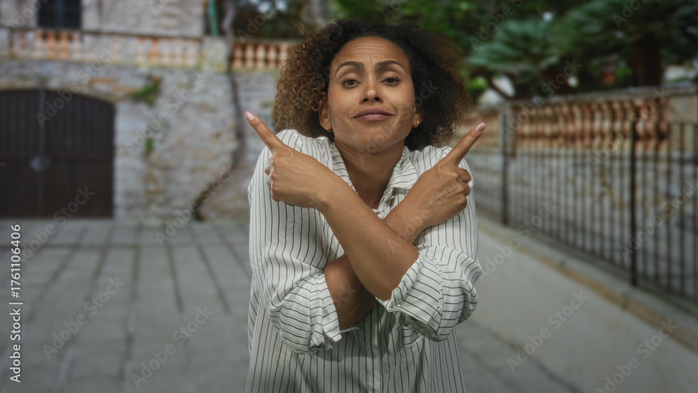 Fototapeta premium Woman with crossed arms forming an x, index fingers pointing opposite ways on a cobblestone street by a stone building; dismissal uncertainty.