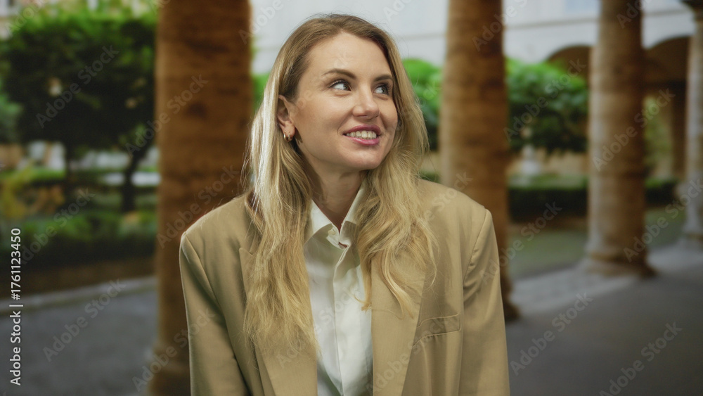 Fototapeta premium Woman smiling in outdoor setting with blonde hair and beige jacket, surrounded by lush green plants and architectural columns on a bright day.