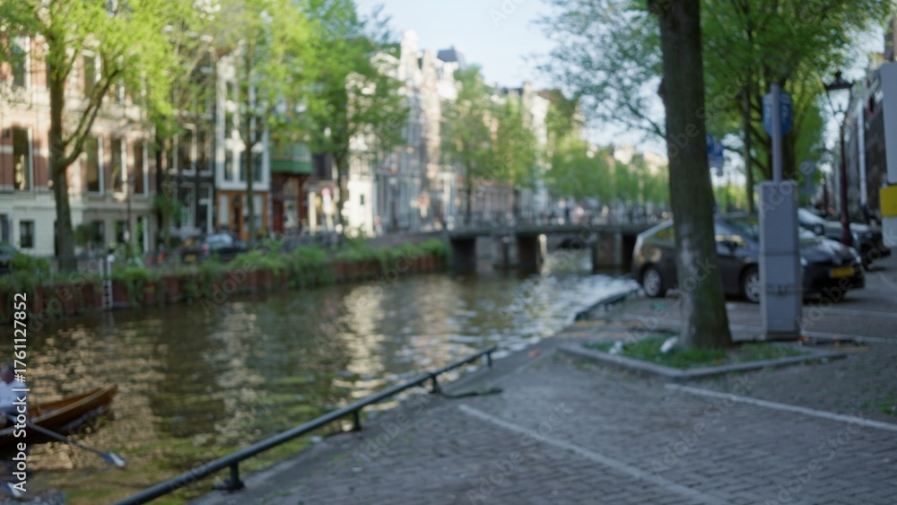 Obraz premium Urban canal scene in amsterdam with blurred buildings and a man rowing in a bokeh backdrop under the greenery of the netherlands.