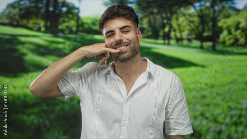 Man smiling makes call gesture with his hand near his face in green sunny park outdoors; communication happiness.