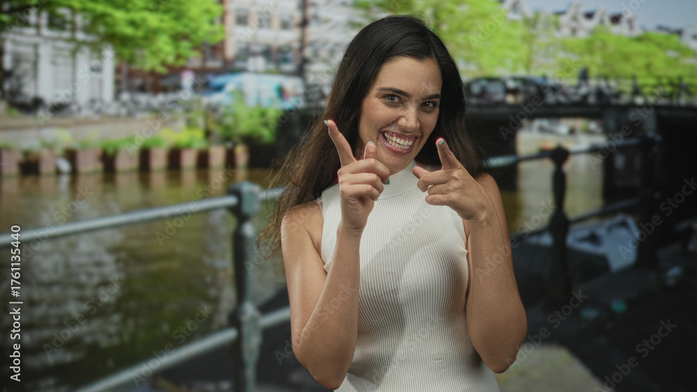 Fototapeta premium Young hispanic woman points finger at canal railing on street in amsterdam city outdoors; confidence boldness empowerment assertiveness.