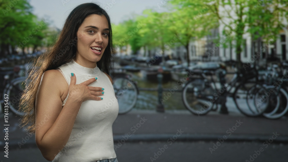 Fototapeta premium Woman smiling and waving on street by an amsterdam canal with bicycles and water in view, wearing a white sleeveless top; gratitude warmth connection calm.