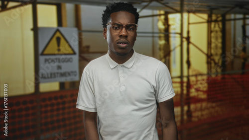 Man touching chin at outdoor construction site with scaffolding and warning sign visible; uncertainty and doubt.