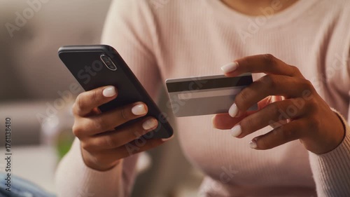 A young African American woman with curly hair sits comfortably at home, using her smartphone while holding a credit card, focused on making an online purchase.