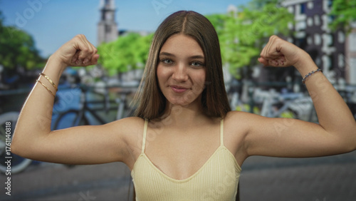 Foto Caucasian teenage girl smiles and flexes arm beside canal on sunlit street amid historic buildings; youth confidence