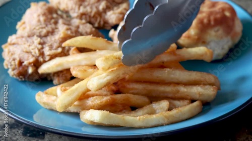 Tongs serving french fries onto a blue plate with crispy fried chicken and a buttery biscuit