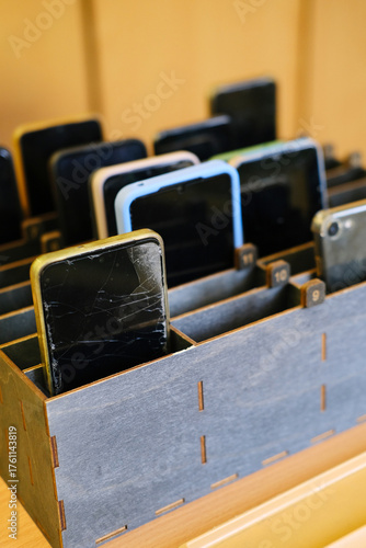 Collection of cell phones stored in a wooden box, showcasing various devices with cracked screens, representing the ban on mobile phones in educational environments and promoting focus in classrooms