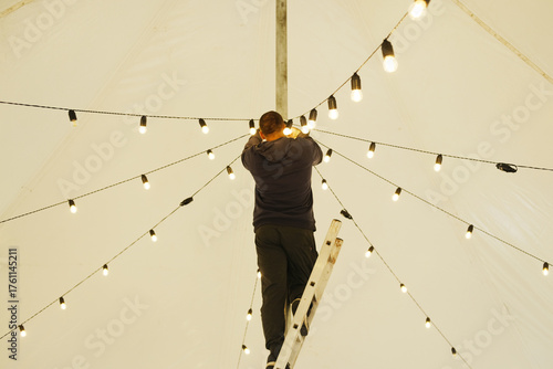 Male technician adjusting decorative string lights on a ladder inside a large tent, creating a warm ambiance for an outdoor event with illuminated atmosphere