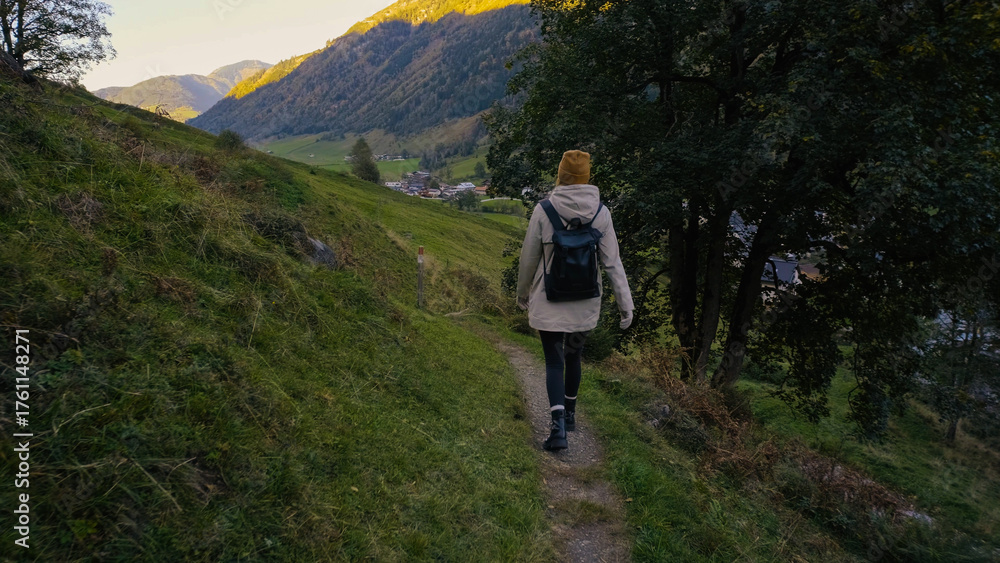 Obraz premium Hiking along a winding path in the mountains during golden hour, surrounded by lush greenery and scenic views of a quaint village