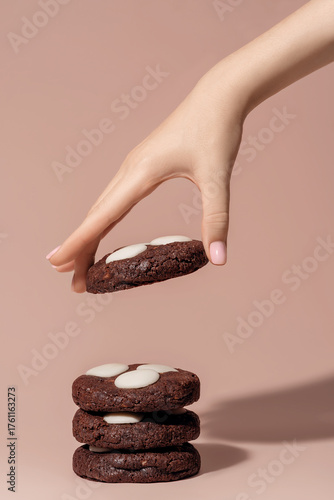 Hand reaching for chocolate cookies with white chocolate chips on a soft pastel background with copy space
