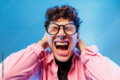 Photos Stressed teen in glasses screaming with hands behind head on blue studio backgro