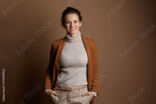 A smiling businesswoman in her 30s-40s, dressed in a brown blazer and beige trousers, stands with hands in pockets, exuding a confident and approachable demeanor in this studio portrait.