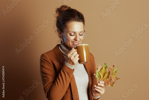 Tableau sur toile A businesswoman, eyes closed in a blissful smile, brings a yellow mug to her lips while holding autumn leaves, exuding cozy contentment against a warm brown autumnal backdrop