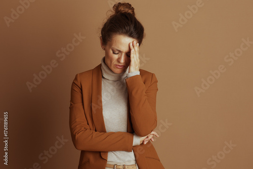 A professional woman, in a brown blazer and beige turtleneck, looks down with a pained expression and hand to her forehead, conveying discomfort, stress, or exhaustion against a brown background.