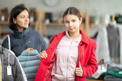 Fototapeta Teen girl is fitting on and trying on red coat in store, family shopping