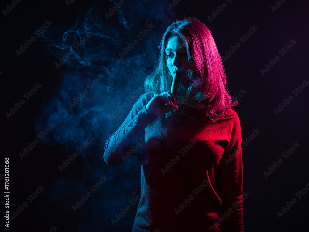 Obraz premium Woman vaper in dark room. Woman inhales vapor from mouth. Young girl smokes vape. Student with vape device in puff of smoke. Concept smoking e-cigarette instead tobacco. Woman vape illuminates light