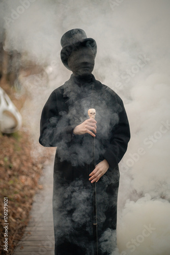 A man with a face in black bandages is dressed in an old vintage style: a classic black coat, a top hat, a walking stick with a skull in his hands. Smoke is coming out of this man and around him. 