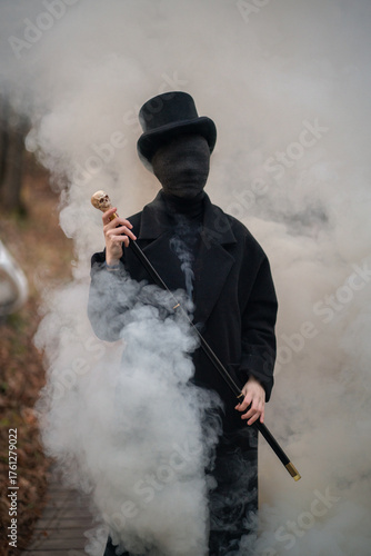 A man with a face in black bandages is dressed in an old vintage style: a classic black coat, a top hat, a walking stick with a skull in his hands. Smoke is coming out of this man and around him. 