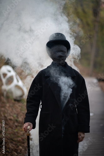 A man with a face in black bandages is dressed in an old vintage style: a classic black coat, a top hat, a walking stick with a skull in his hands. Smoke is coming out of this man and around him. 