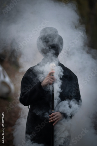 A man with a face in black bandages is dressed in an old vintage style: a classic black coat, a top hat, a walking stick with a skull in his hands. Smoke is coming out of this man and around him. 