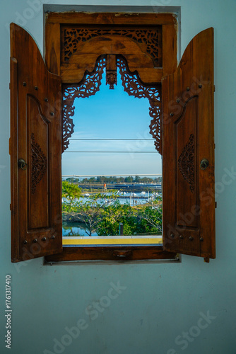 Wooden windows with carved motifs that open to reveal the outside view of water, trees and bright blue sky.