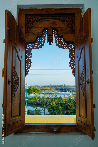 Wooden windows with carved motifs that open to reveal the outside view of water, trees and bright blue sky.