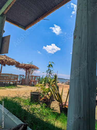 a view of a bright blue sky on a sandy beach with a few wooden chairs and straw umbrellas from behind the window frame.