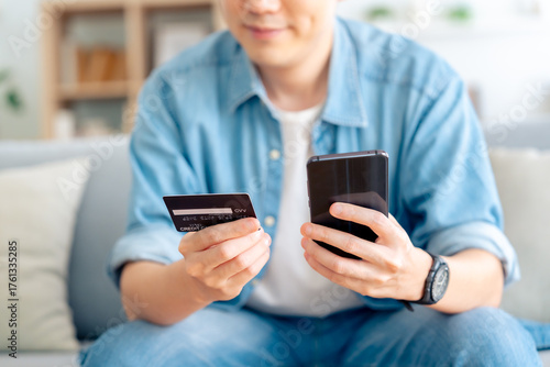 Young asian man sitting on sofa in living room, makes online banking payments through the internet from bank card on cell phone. Shopping online on mobile with credit card