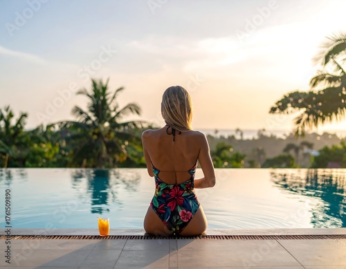 A woman in a floral swimsuit sits at an infinity pool's edge, facing a vibrant sunset. Tropical trees frame the serene scene with an orange drink nearby