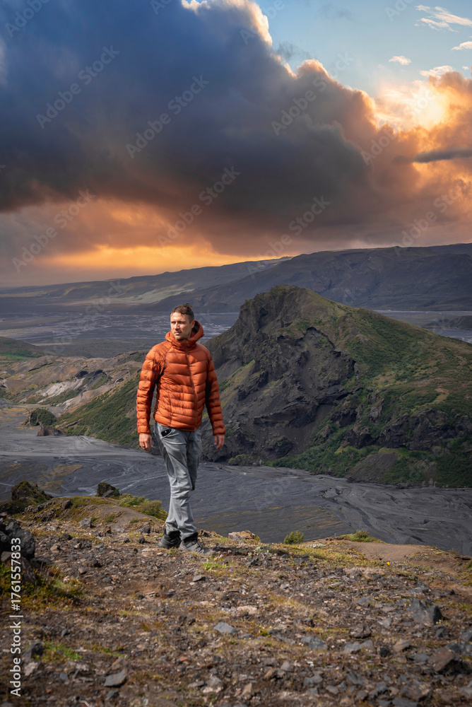 Fototapeta premium A lone hiker walks a rocky ridge at sunset in Iceland highlands. Storm clouds glow above mossy lava slopes and a gray riverbed near a craggy peak, with rich contrast.