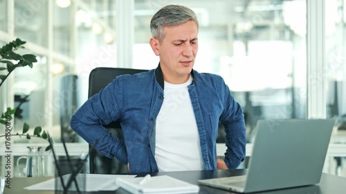 Stressed businessman in an office experiences back pain while working on laptop, wearing a denim jacket. Pain from prolonged sitting and computer use.