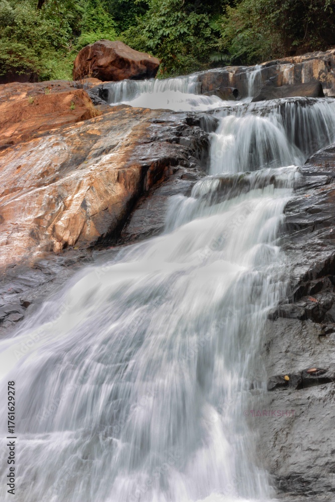 Obraz premium Soft flowing waterfall cascading over rocky steps in a forest setting. The smooth water effect contrasts with textured rocks and lush greenery, capturing a tranquil natural scene.