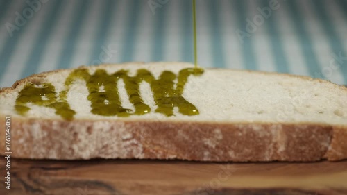 Close-up of a steady stream of extra virgin olive oil dripping onto a slice of artisan bread on a wooden board.