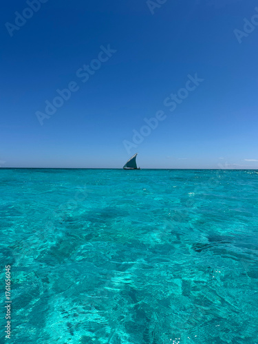 A vast, sparkling turquoise ocean stretches toward the horizon in Madagascar, with a small sailboat visible in the distance. The bright sunlight reflects off the calm water, creating a serene and idyl
