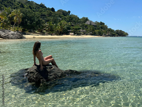 A woman sitting on a rock in the shallow waters near Nosy Komba island, Madagascar, surrounded by a pristine beach, white sand, and crystal-clear turquoise ocean. The bright sunlight and serene tropic