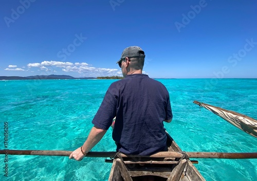 A man paddling a wooden boat toward a tropical island in Madagascar across clear turquoise waters. The bright sunlight reflects on the calm ocean, highlighting the white sandy shores and creating a pe