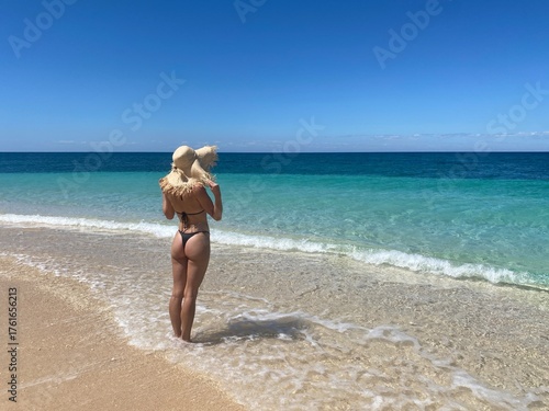 A woman standing on a beautiful tropical beach in Madagascar with clear turquoise water and white sand. The bright sunlight, blue sky, and crystal-clear ocean create a peaceful and exotic summer 