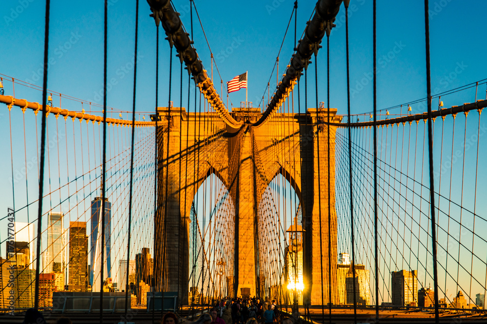 Fototapeta premium Iconic Brooklyn Bridge Towers Bathed in Golden Sunrise Light, Featuring American Flag and New York City Skyline