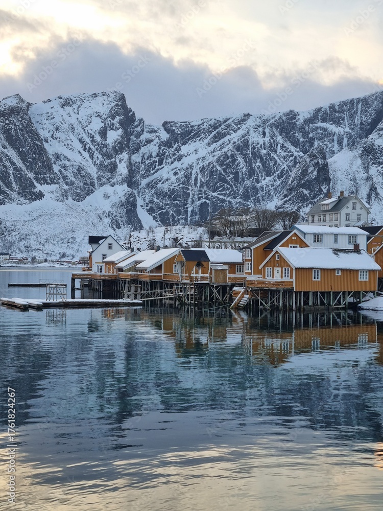 Fototapeta premium Sakrisoy fishing village and snowy mountains in winter on Lofoten islands, Norway. Winter landscape with traditional yellow rorbu houses on stilts in fjord water. Seascape of Norwegian sea. 