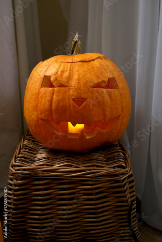 Рumpkin with a candle in the middle on a wicker stand made of vines on a background of fabrics. A scary face is carved on the pumpkin. Halloween theme, holiday decorations, decoration, celebration