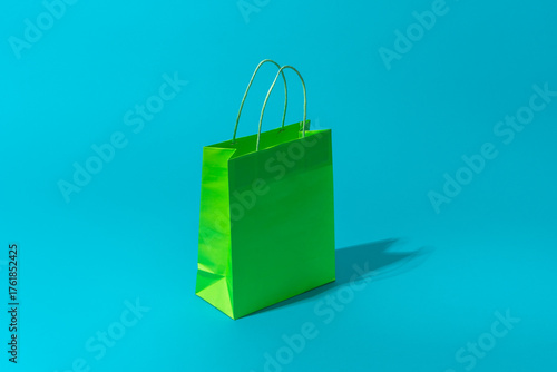 Shopping bag standing on the turquoise blue background with copy space. Minimalist photo of green paper bag with hard shadow central composition. Summer sale and shopping concepts.