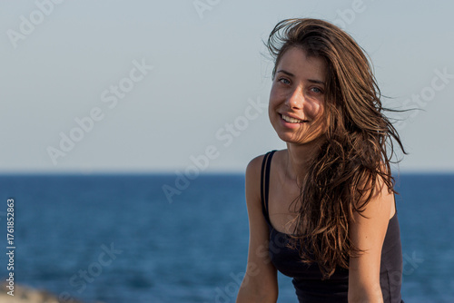 Portrait of cheerful suntanned young woman with sea and blue sky in background. Photo of beautiful brunette girl relaxing on the resort under the hot sun. Summer and vacation concept.