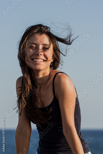 Photo of beautiful brunette girl relaxing on the resort under the hot sun. Summer and vacation concept. Portrait of cheerful suntanned young woman with sea and blue sky in background.