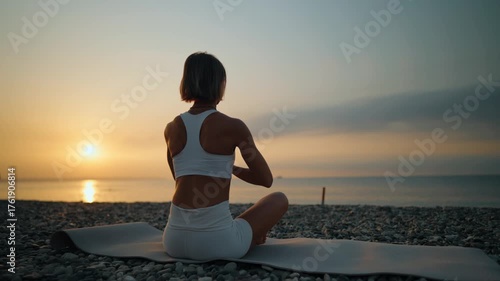 Young woman practices yoga on a mat by the sea during a peaceful morning sunrise. She sits in a meditative posture facing the water. Concept of mindfulness, inner balance, and connection with nature