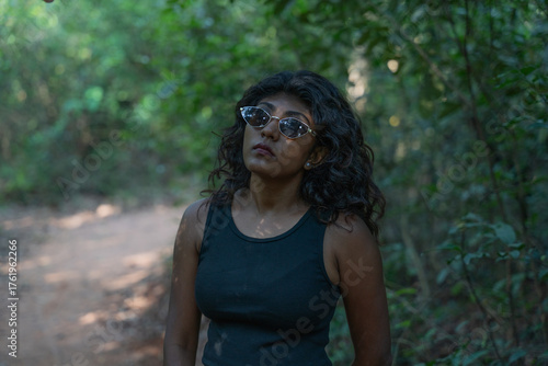 A woman wearing sunglasses and a black tank top stands in a forest