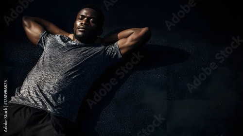 A fit Black man lies on the ground with hands behind his head resting and recovering after an outdoor workout bathed in sunlight and shadow